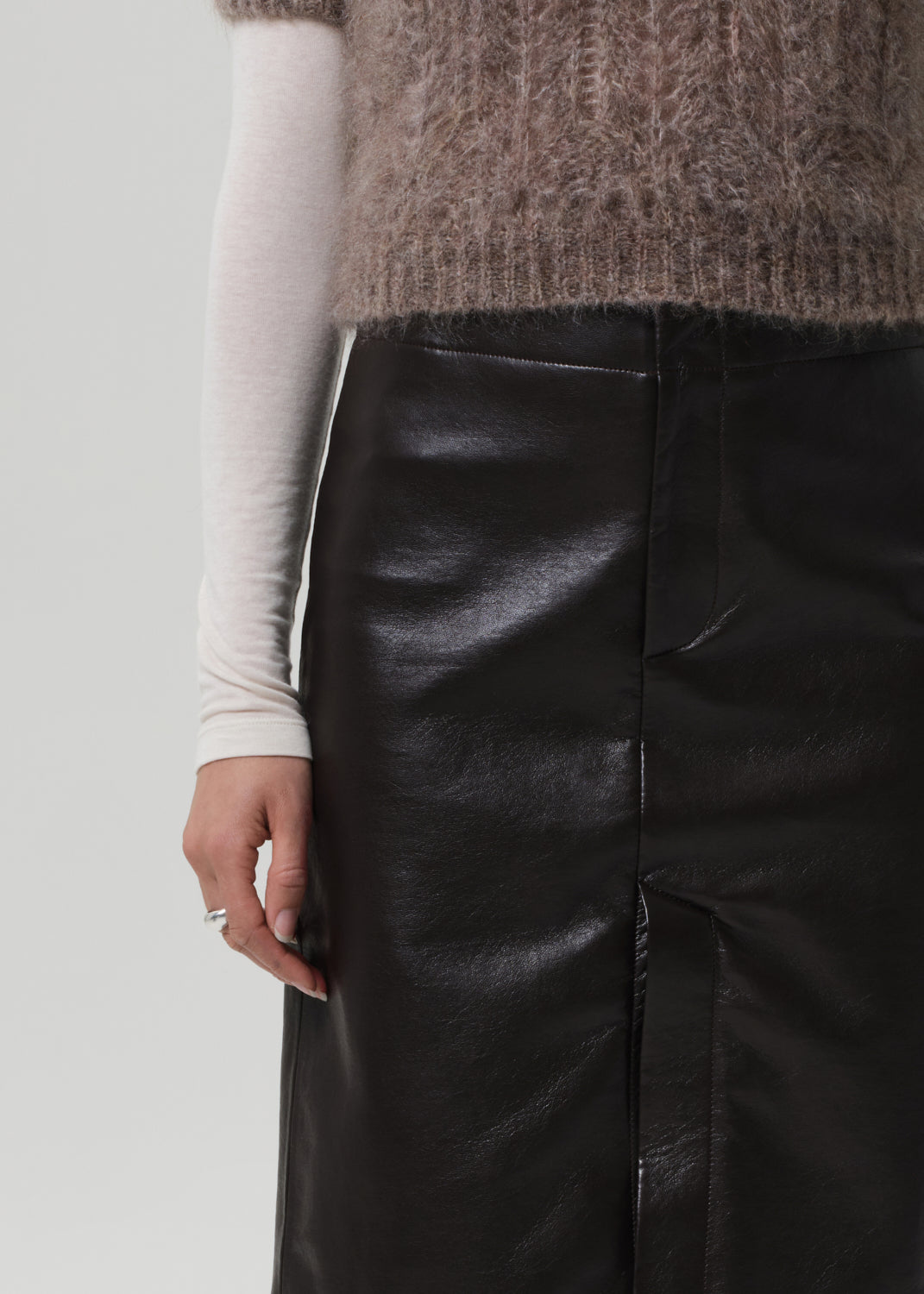 Person wearing a brown knitted sweater, brown leather skirt, and black knee-high boots on a grey background