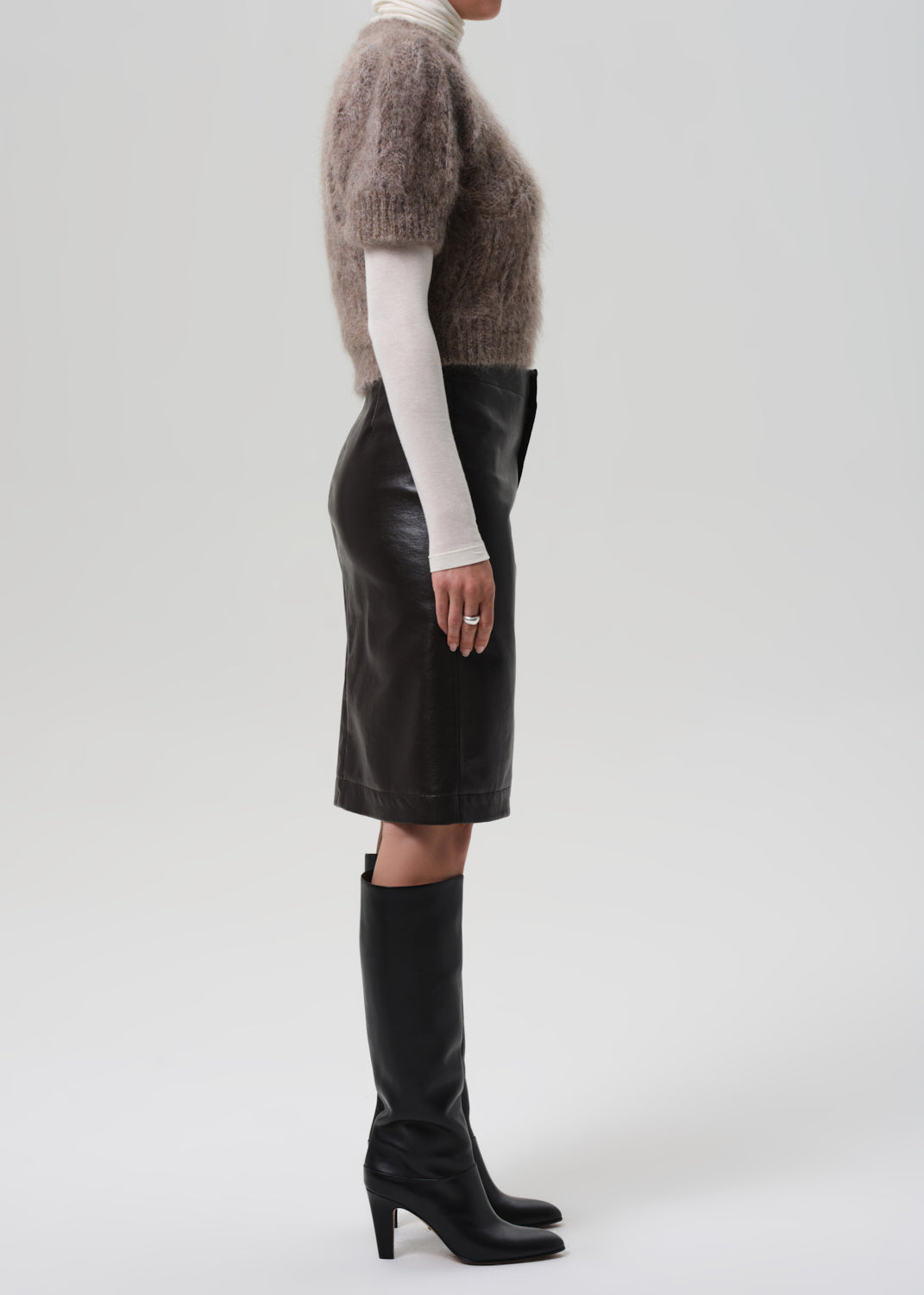 Person wearing a brown knitted sweater, brown leather skirt, and black knee-high boots on a grey background