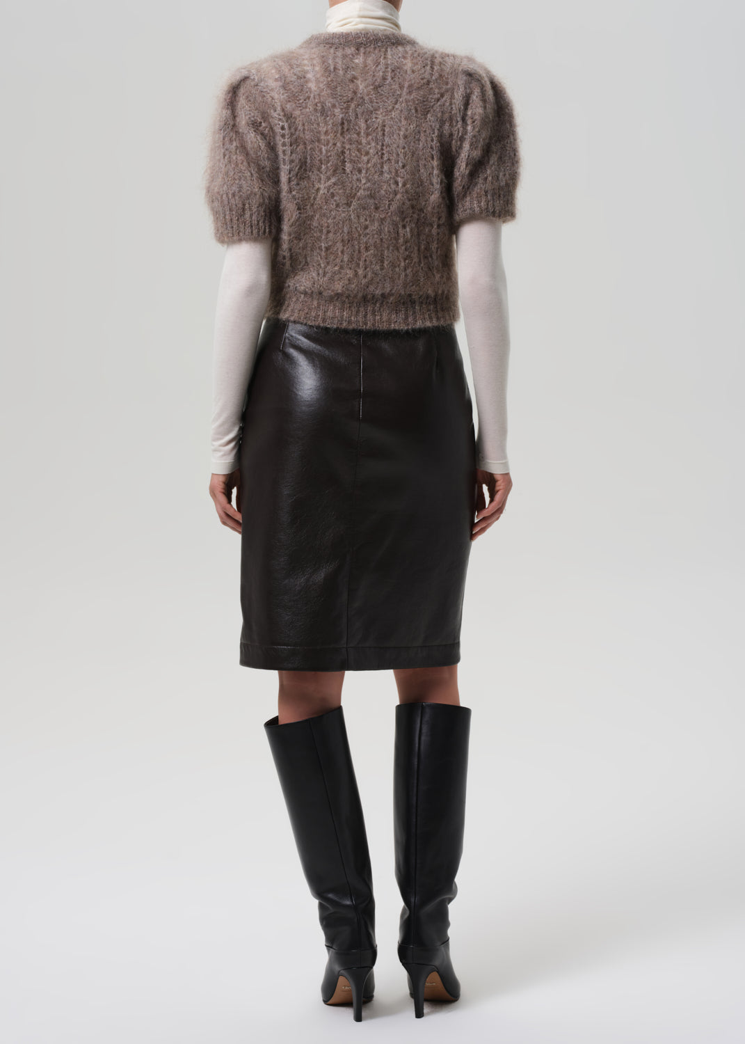 Person wearing a brown knitted sweater, brown leather skirt, and black knee-high boots on a grey background