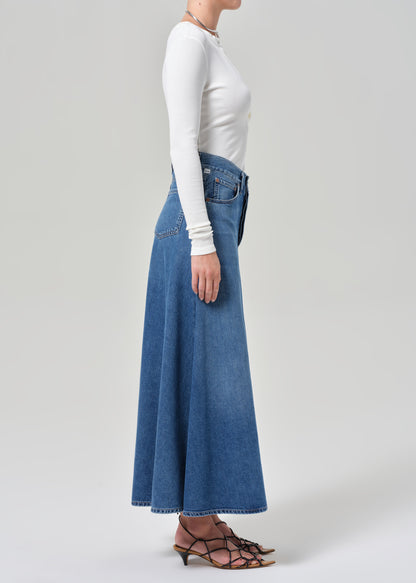 Person wearing a long blue denim skirt and white top on a plain background