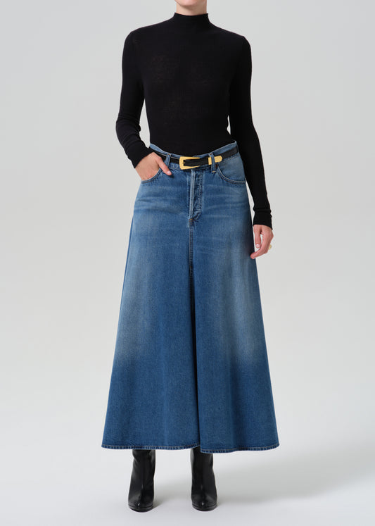 A model wearing a black long-sleeve mock neck top and a high-waisted, wide-leg blue denim skirt with a yellow-gold belt, paired with black ankle boots, against a light grey background.