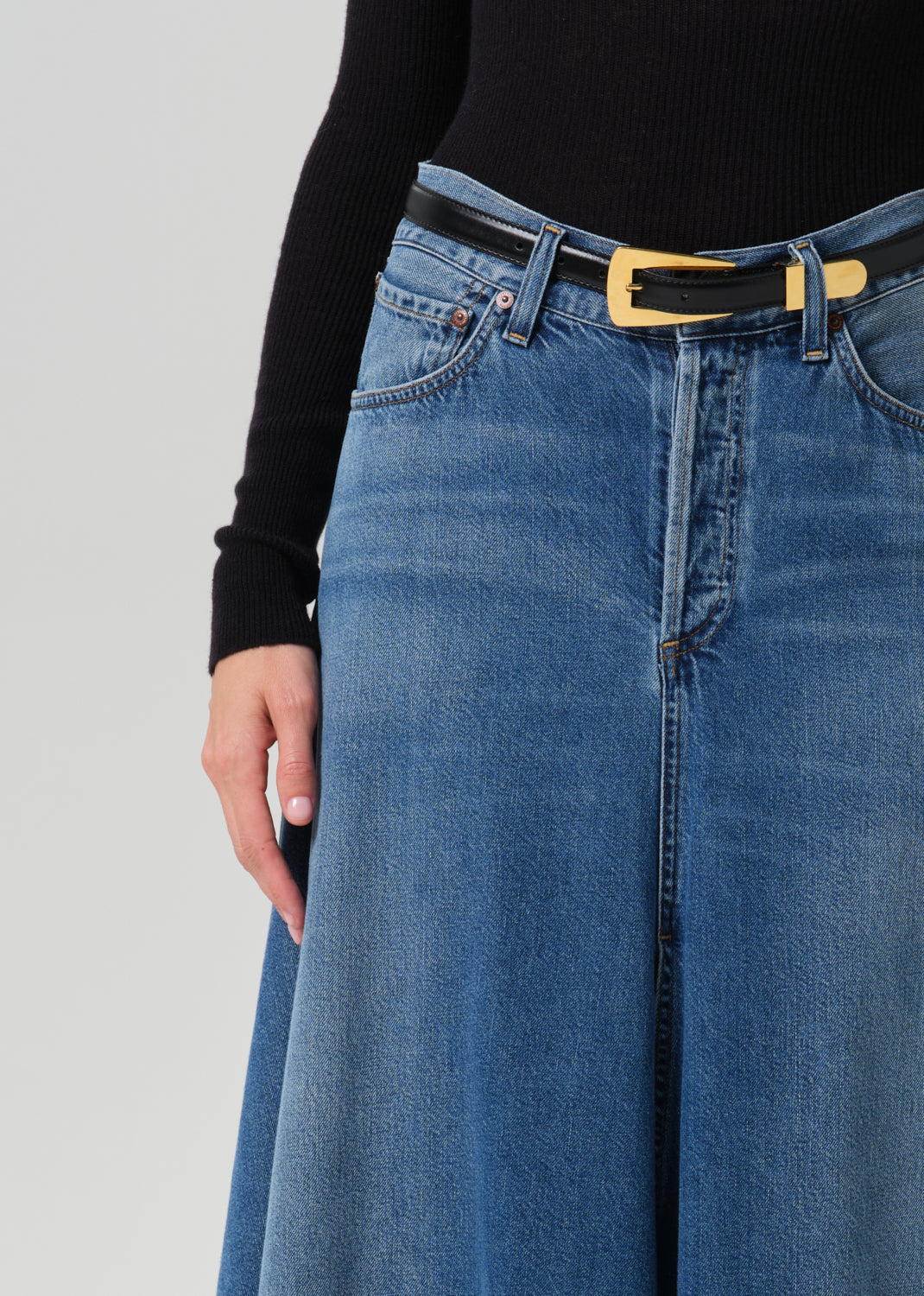 Close-up of a person wearing a blue wide-leg denim skirt with a black belt featuring a gold rectangular buckle, paired with a black long-sleeve top, against a grey background.