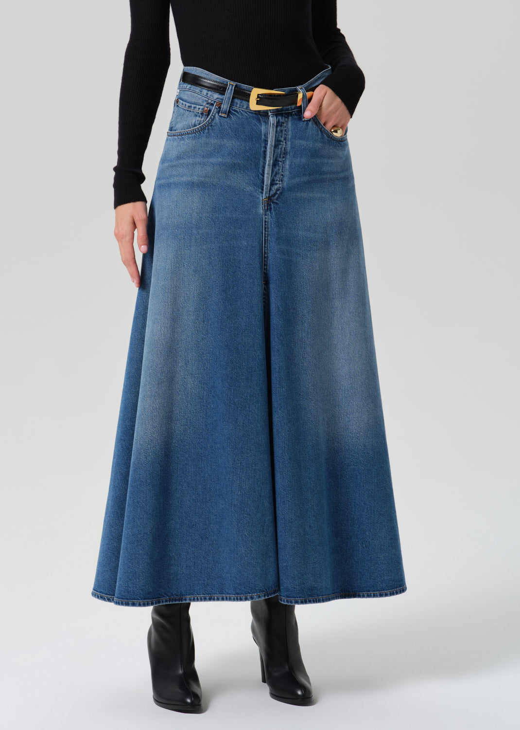 A model wearing a high-waisted, wide-leg blue denim skirt with a black belt and gold buckle, styled with a black long-sleeve top and black ankle boots against a grey background.