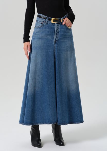 A model wearing a high-waisted, wide-leg blue denim skirt with a black belt and gold buckle, styled with a black long-sleeve top and black ankle boots against a grey background.