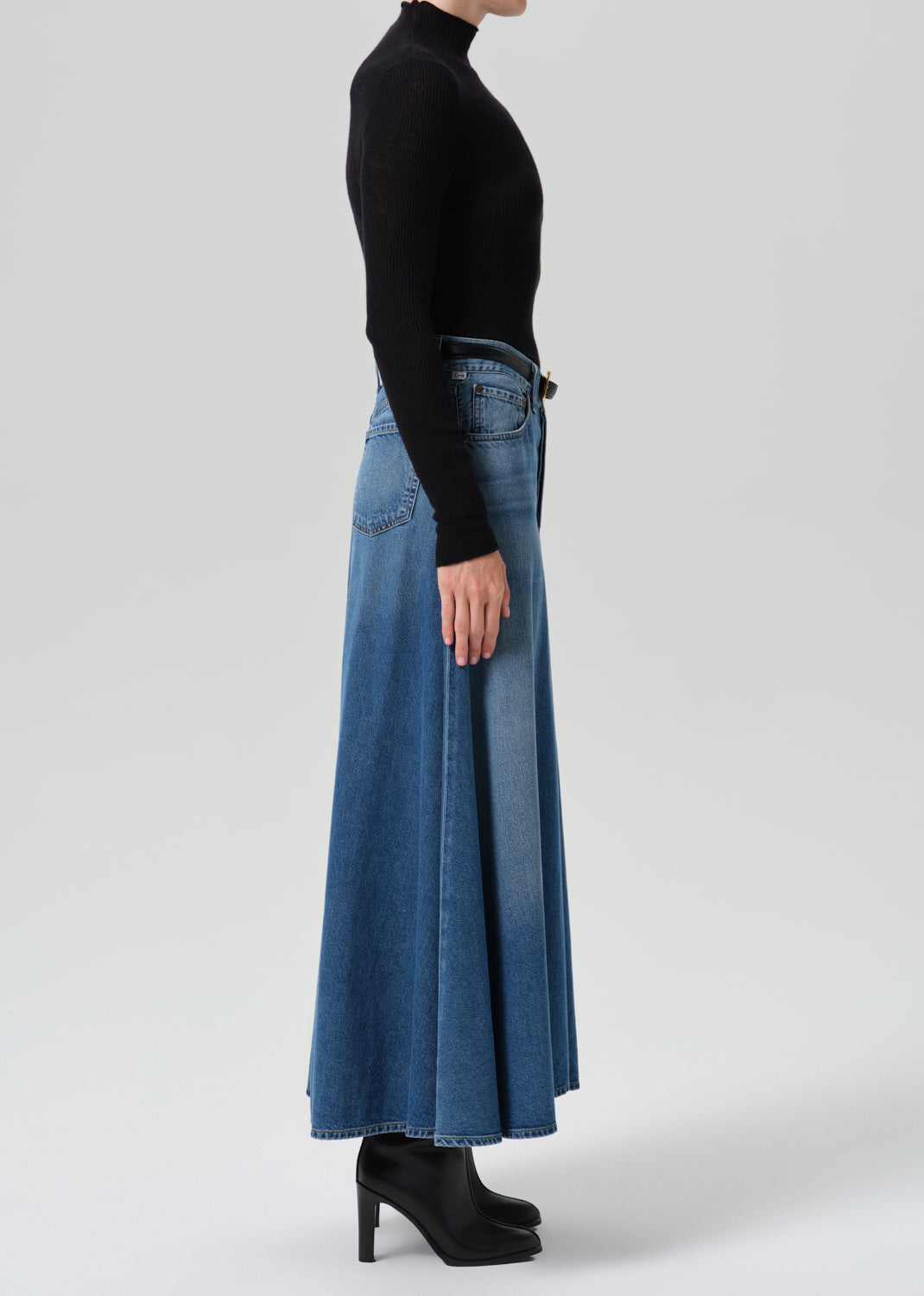A model shown from the side wearing a black long-sleeve mock neck top and a high-waisted, wide-leg blue denim skirt with black high-heeled boots, against a grey background.