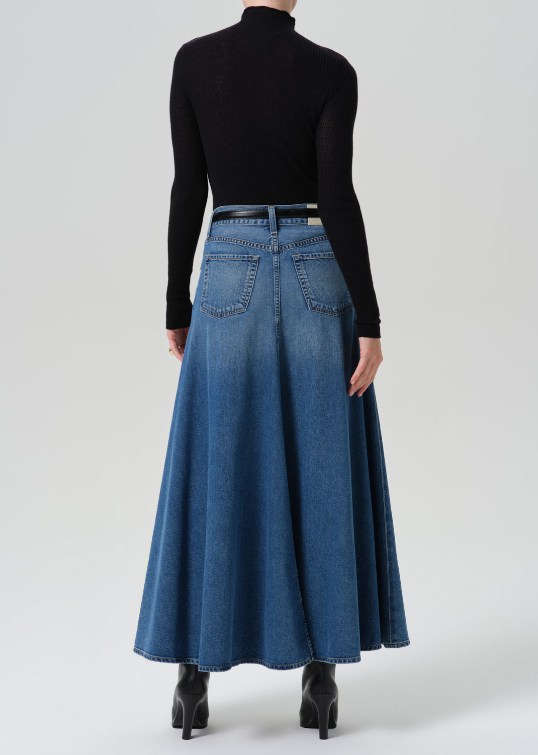 A model shown from the back wearing a long-sleeve black mock neck top and a high-waisted, flared blue denim maxi skirt with a thin black belt, paired with black ankle boots, against a grey background.