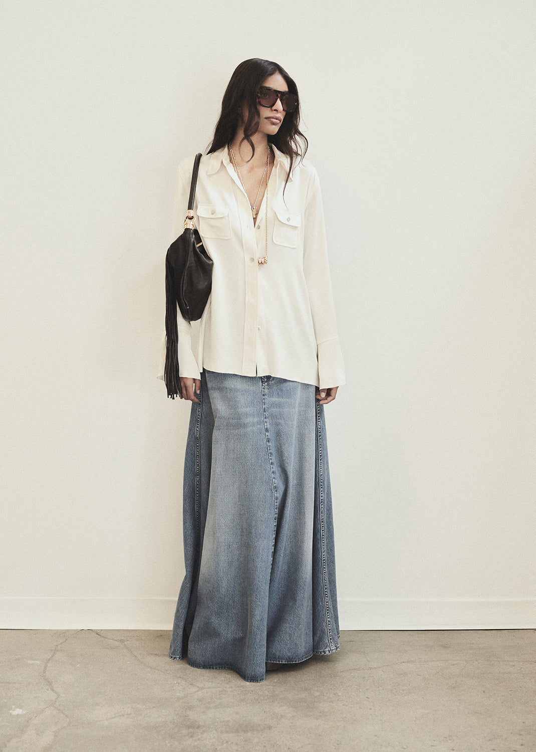 Woman wearing a white blouse and denim skirt with sunglasses and a black bag against a white background