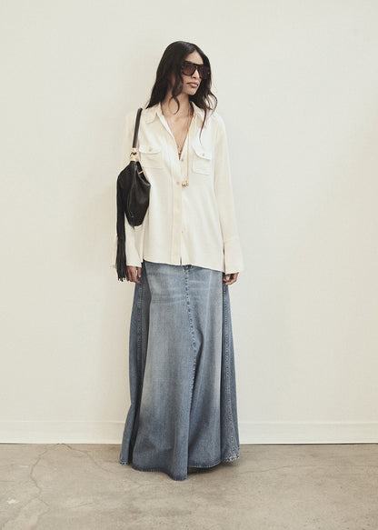 Woman wearing a white blouse and denim skirt with sunglasses and a black bag against a white background