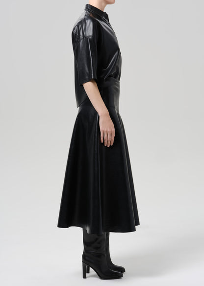 Person wearing a black leather short sleeve shirt with a black leather midi skirt viewed from the side on a grey background