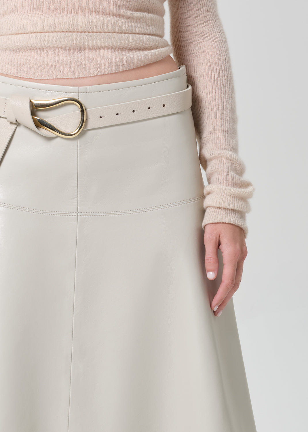 Person wearing a light pink sweater and light grey leathers skirt with a belt on a grey background