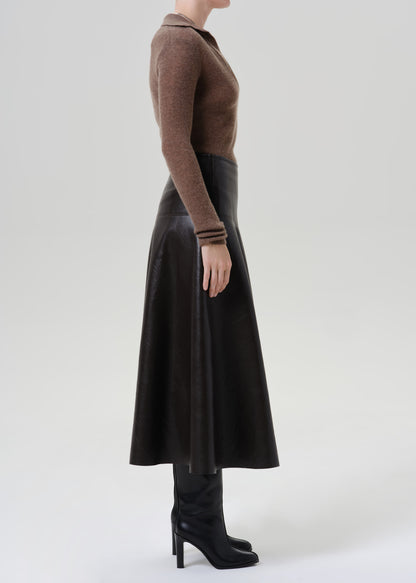 Person wearing a brown long sleeve collared knit top with a brown leather midi skirt viewed from the side on a grey background