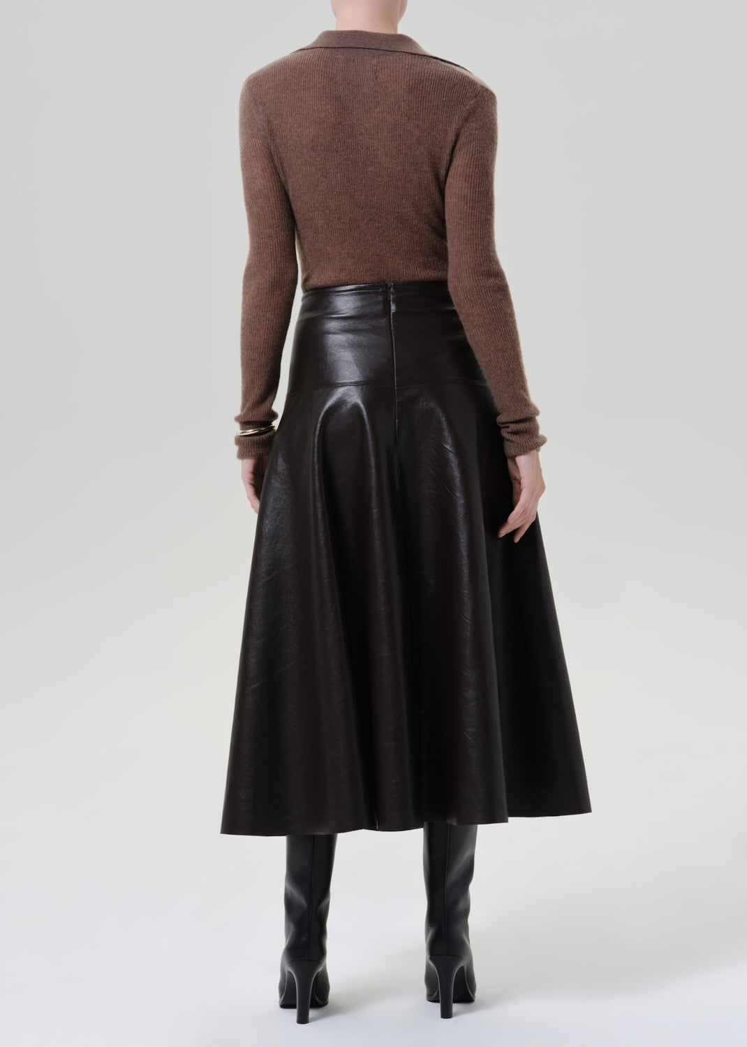 Person wearing a brown long sleeve collared knit top with a brown leather midi skirt viewed from the back on a grey background