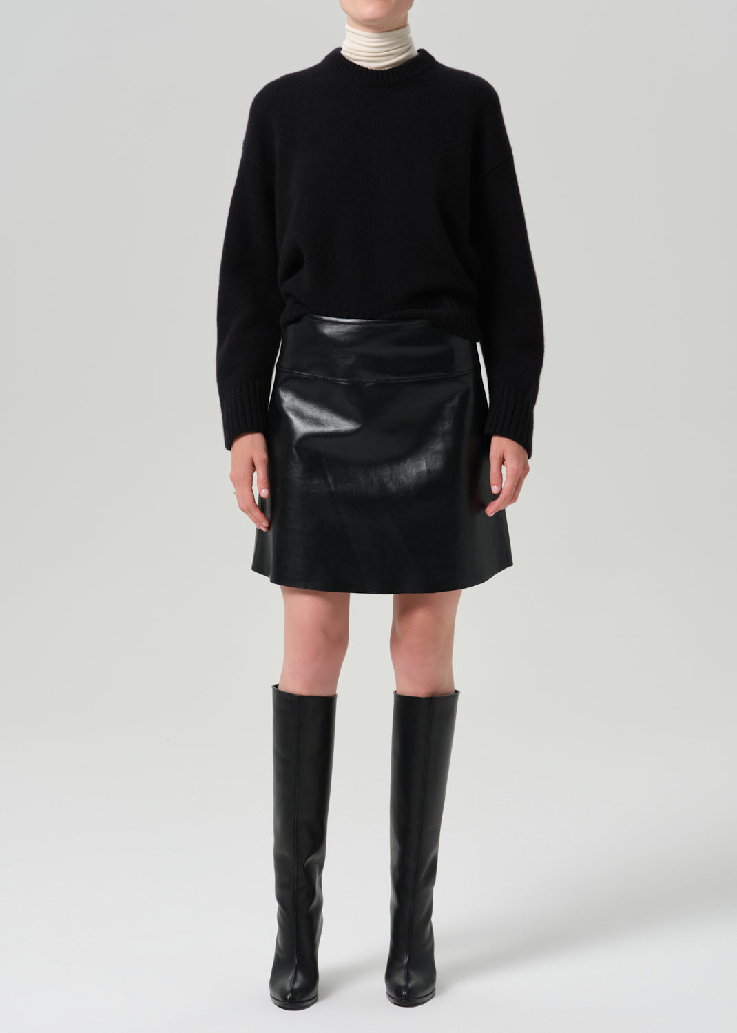 A model wearing a black knit mock-neck sweater, a black faux-leather mini skirt, and black leather knee-high boots, standing against a grey background.