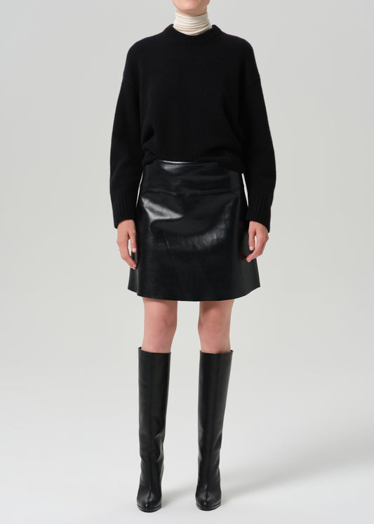 A model wearing a black knit mock-neck sweater, a black faux-leather mini skirt, and black leather knee-high boots, standing against a grey background.