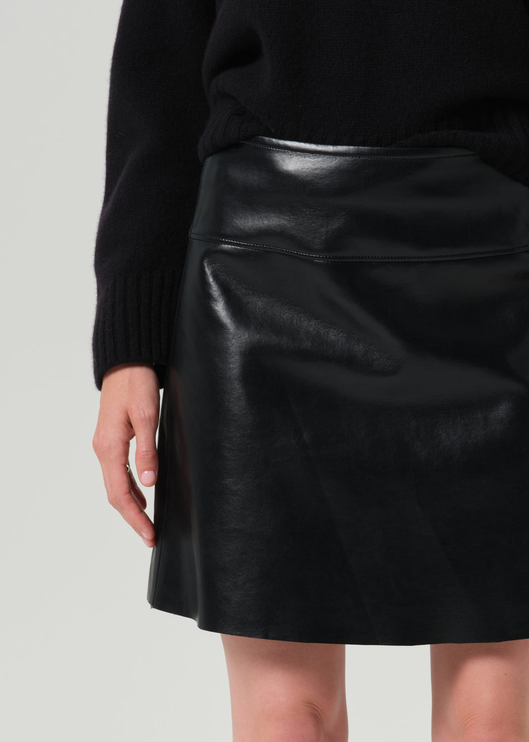 A close-up of a person wearing a black faux-leather mini skirt and a black ribbed sweater against a grey background.