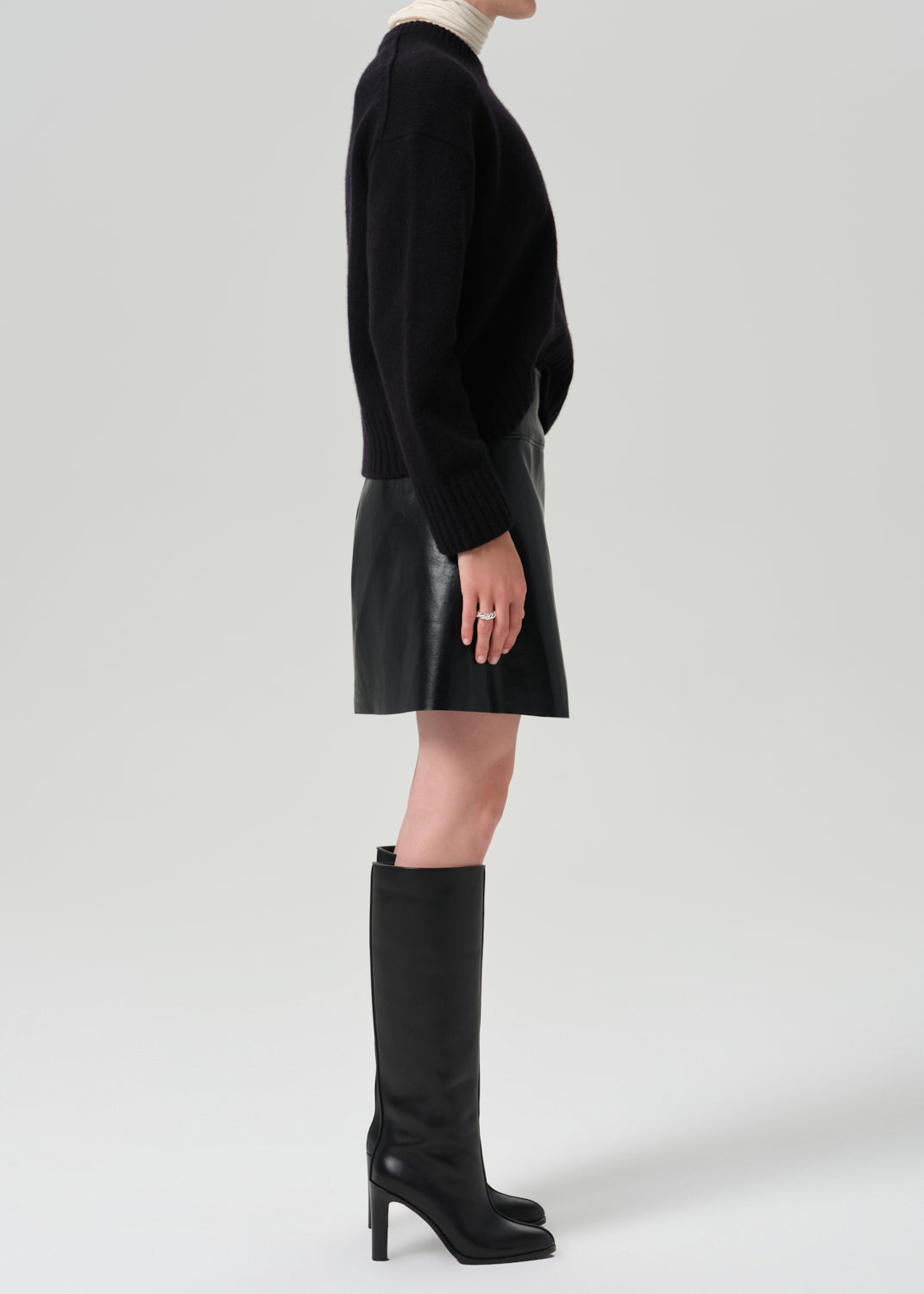 A model shown in profile wearing a black knit sweater, a black leather mini skirt, and black knee-high heeled boots against a grey background.