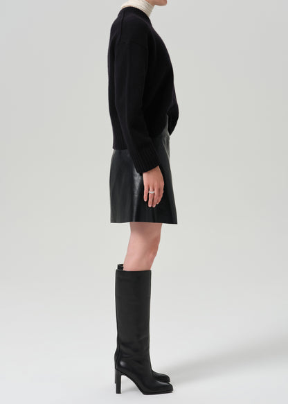 A model shown in profile wearing a black knit sweater, a black leather mini skirt, and black knee-high heeled boots against a grey background.