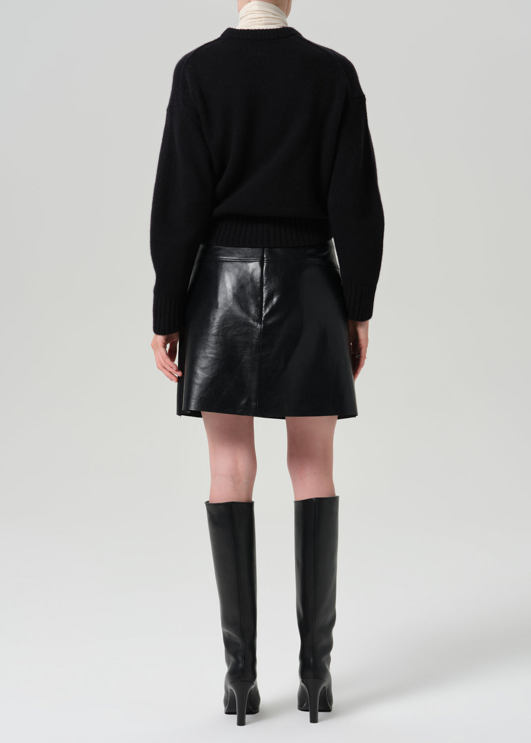A model shown from the back wearing a black knit sweater, a black faux-leather mini skirt, and black knee-high heeled boots against a grey background.