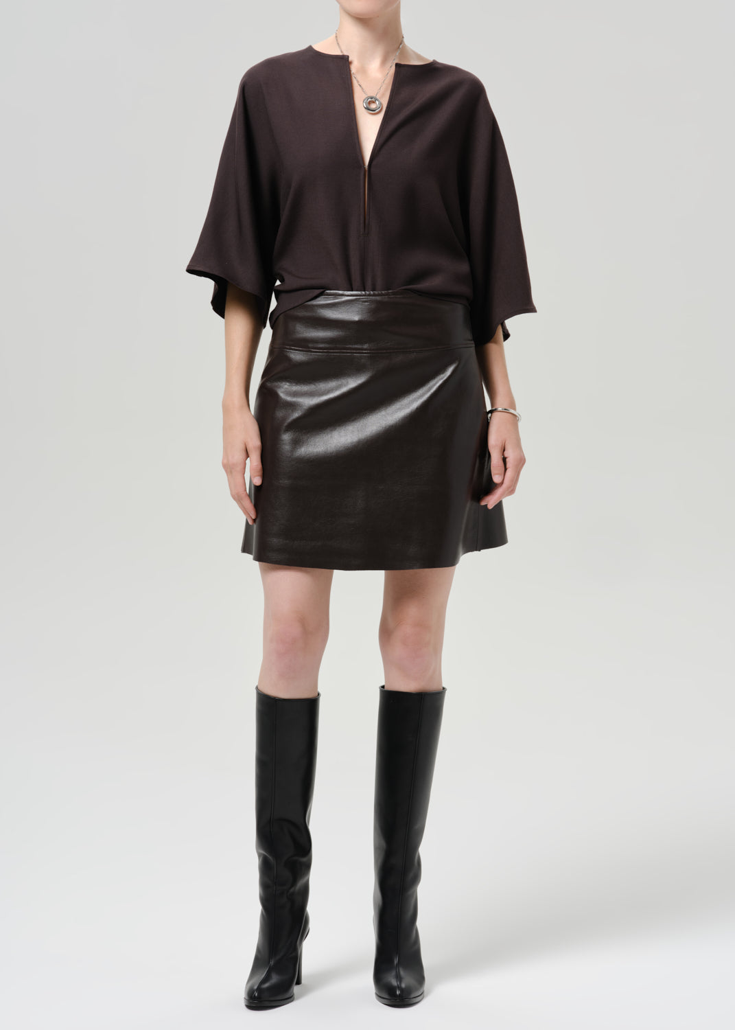 Person wearing a dark brown leather mini skirt with a dark brown top and black knee high boots on a grey background