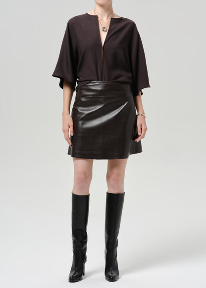 Person wearing a dark brown leather mini skirt with a dark brown top and black knee high boots on a grey background