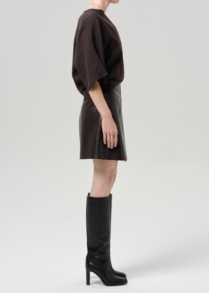 Person wearing a dark brown leather mini skirt with a dark brown top and black knee high boots viewed from the side on a grey background
