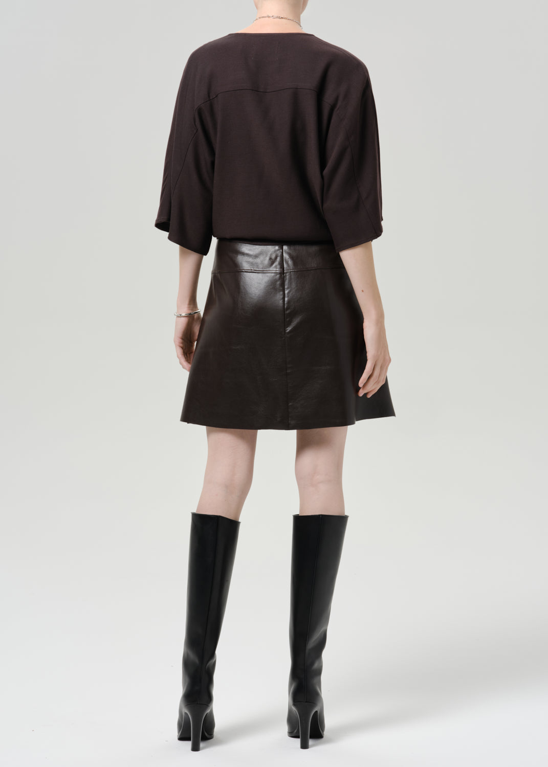Person wearing a dark brown leather mini skirt with a dark brown top and black knee high boots viewed from the back on a grey background