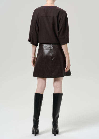 Person wearing a dark brown leather mini skirt with a dark brown top and black knee high boots viewed from the back on a grey background