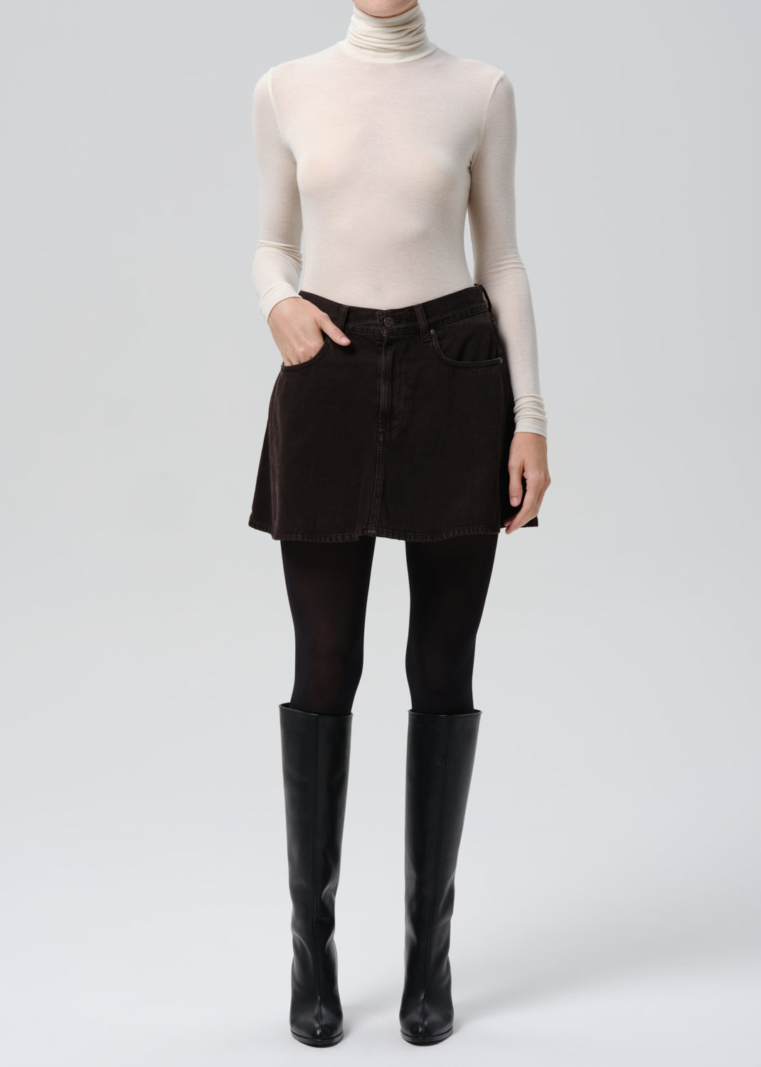 Person wearing a beige turtleneck, black pants, and black knee-high boots on a grey background