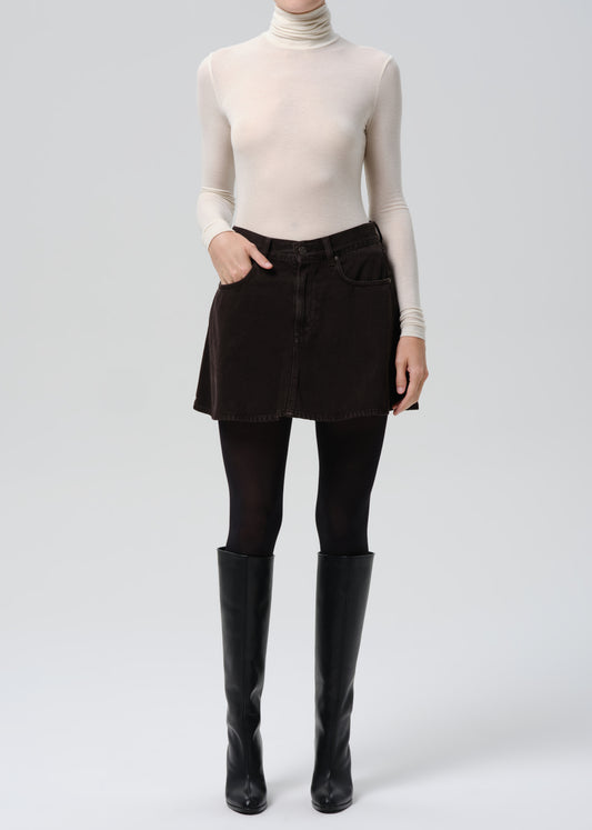 Person wearing a beige turtleneck, black pants, and black knee-high boots on a grey background