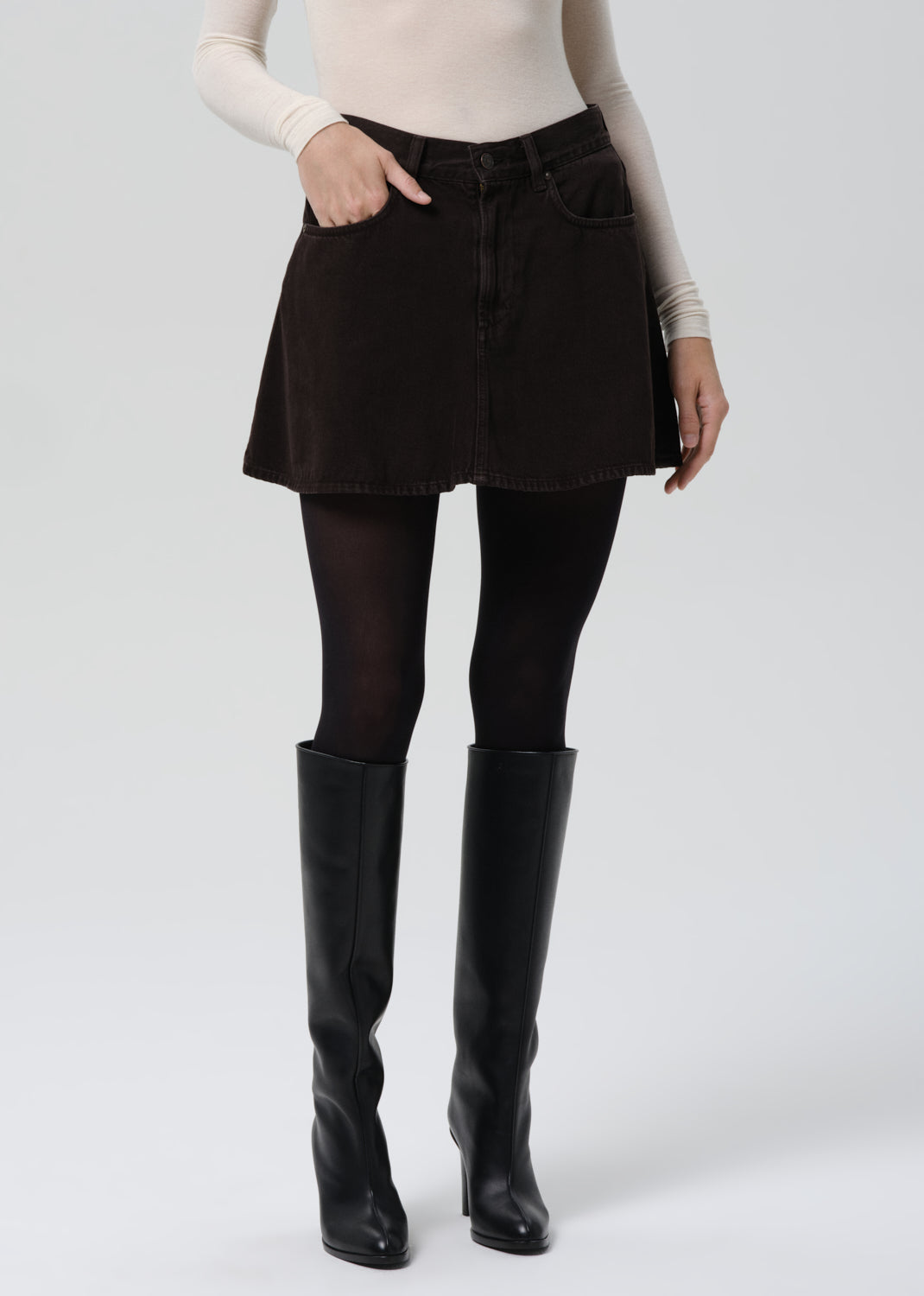 Person wearing a black skirt, black tights, and black knee-high boots on a grey background