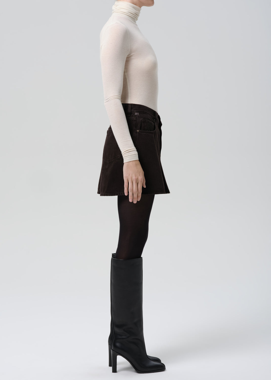 Person wearing a white turtleneck, black skirt, and black knee-high boots on a grey background