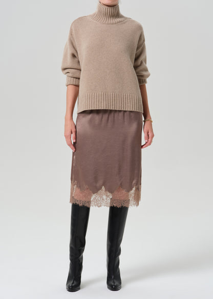 A model wearing a beige turtleneck sweater, a brown satin midi skirt with a lace hem, and black knee-high boots, photographed against a grey background.