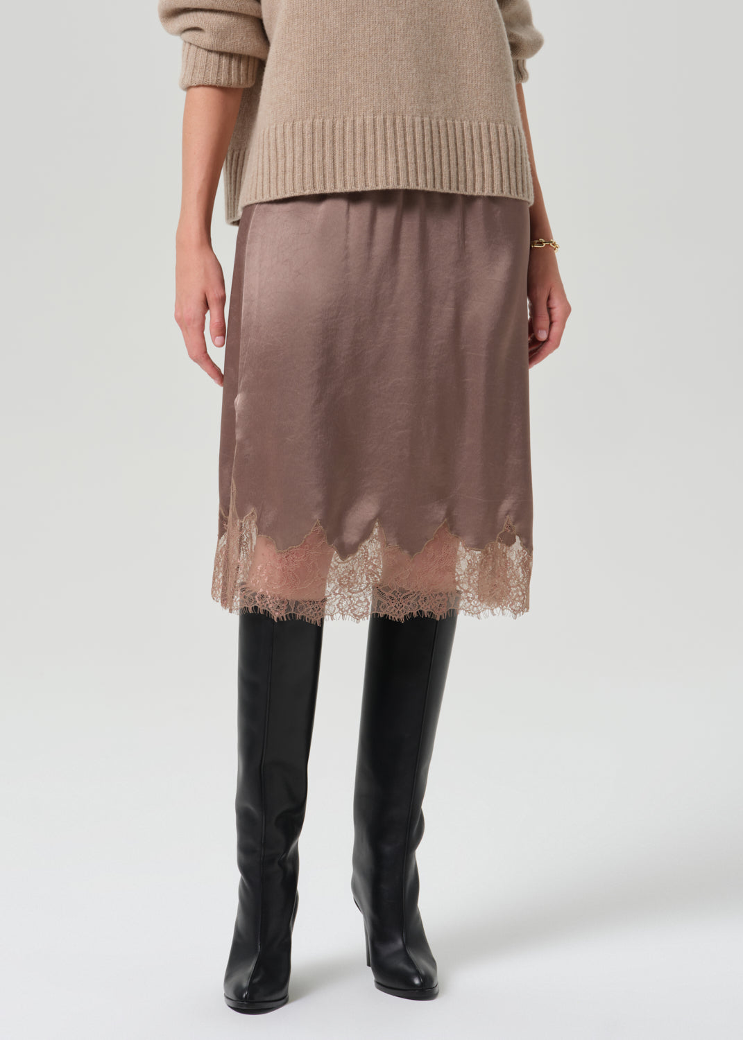 A model is shown from the waist down wearing a taupe brown satin midi skirt with a scalloped lace hem, styled with a beige knit sweater and black knee-high boots against a grey  background.