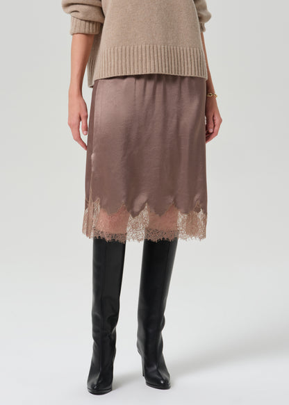 A model is shown from the waist down wearing a taupe brown satin midi skirt with a scalloped lace hem, styled with a beige knit sweater and black knee-high boots against a grey  background.