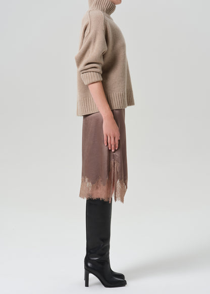 A model shown from the side wearing a beige knit turtleneck sweater, a brown sheer midi skirt with a distressed hem, and black knee-high leather boots, set against a grey background.