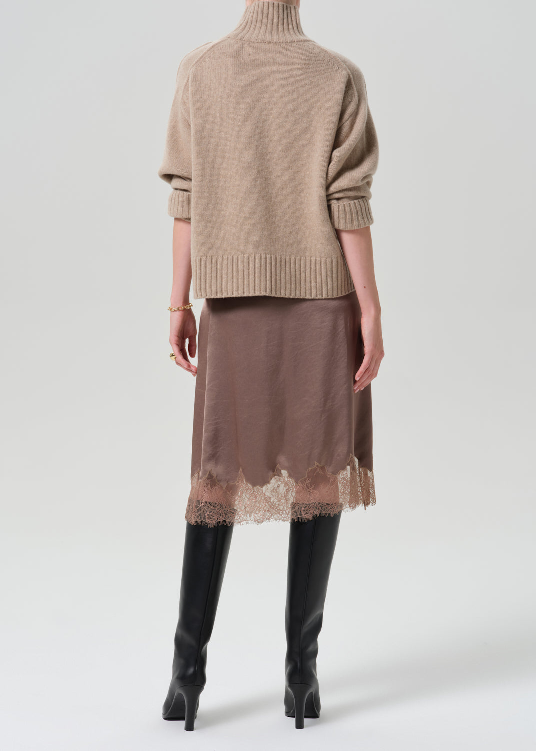 A model shown from the back wearing a beige ribbed turtleneck sweater, a brown midi skirt with a lace hem, and black knee-high boots, against a light grey background.