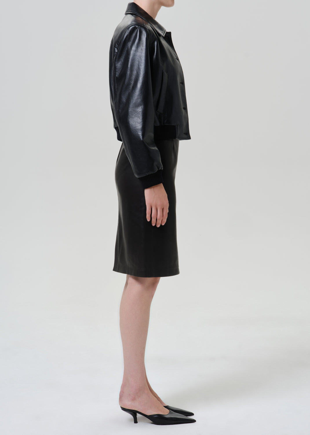 Woman wearing a black leather pencil skirt with a black leather jacket viewed from the side on a grey background.