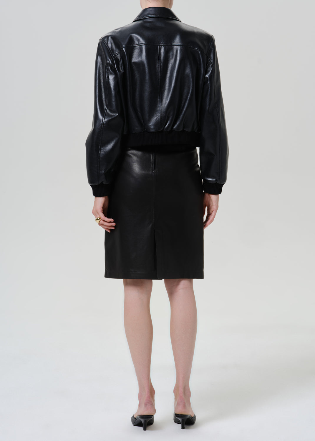 Woman wearing a black leather pencil skirt with a black leather jacket viewed from the back on a grey background.