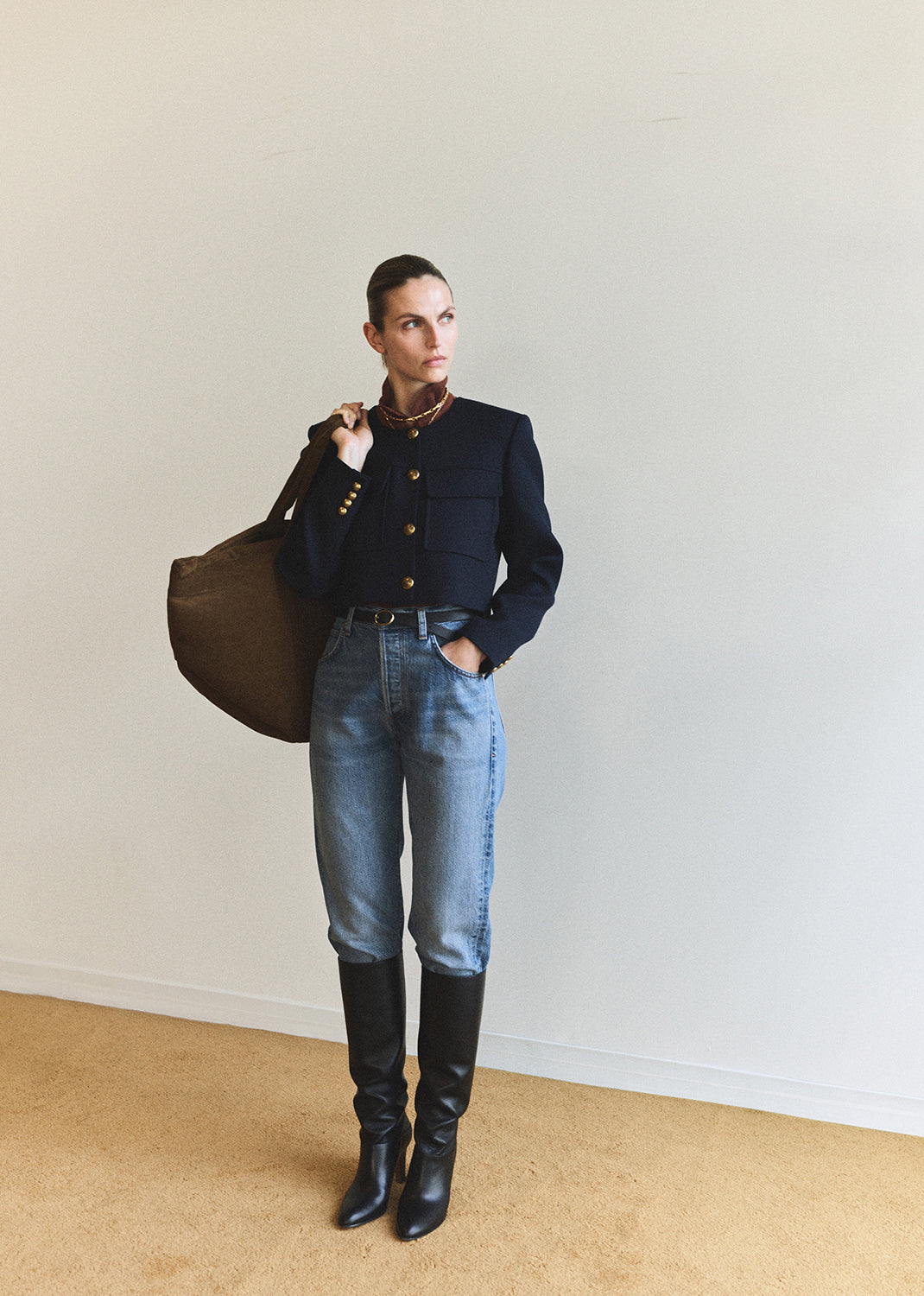 Woman in a navy sweater, jeans, and black boots holding a brown bag against a plain background