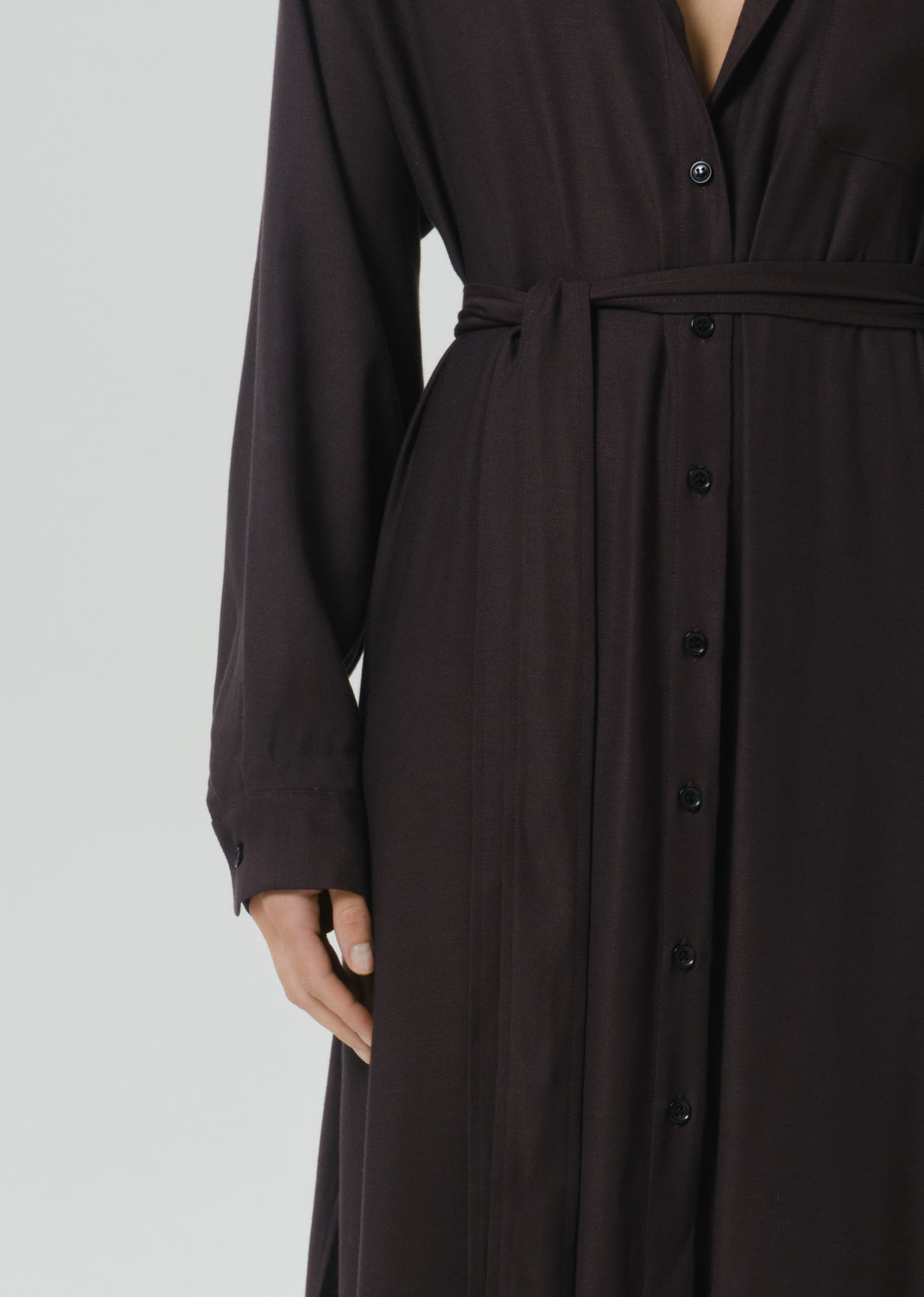 A close-up of a dark brown long-sleeve button-front dress with a matching waist tie, shown on a model against a grey background.