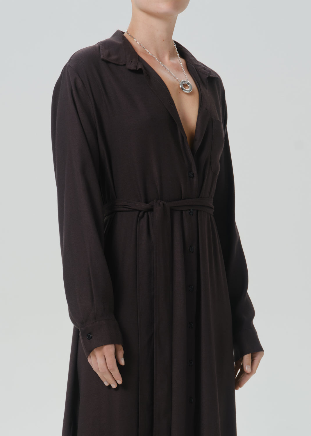 A model wearing a dark brown long-sleeve button-down shirt dress with a tie waist belt, shown from the neck down against a grey background.