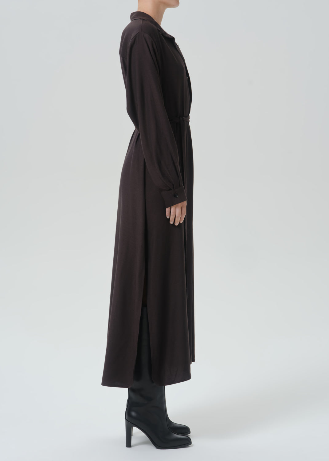 A model shown in profile wearing a long, dark brown button-up shirt dress with a matching waist tie, paired with black high-heeled boots, against a grey background.