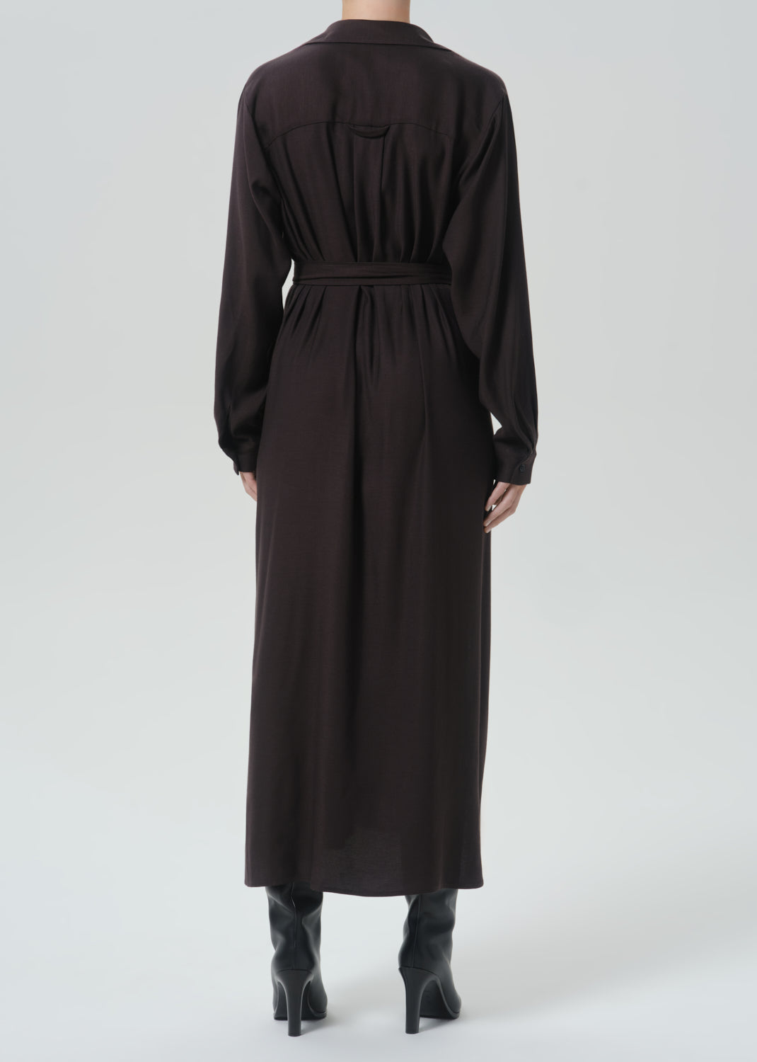 A model viewed from the back wearing a long-sleeved, dark brown midi shirt dress with a matching waist belt, paired with black pointed-toe heeled boots against a grey background.
