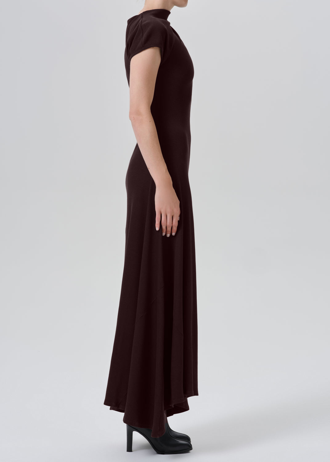 A model shown from the side wearing a dark brown, short-sleeved mock-neck maxi dress with black high heels, set against a grey background.