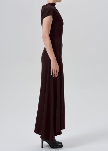 A model shown from the side wearing a dark brown, short-sleeved mock-neck maxi dress with black high heels, set against a grey background.
