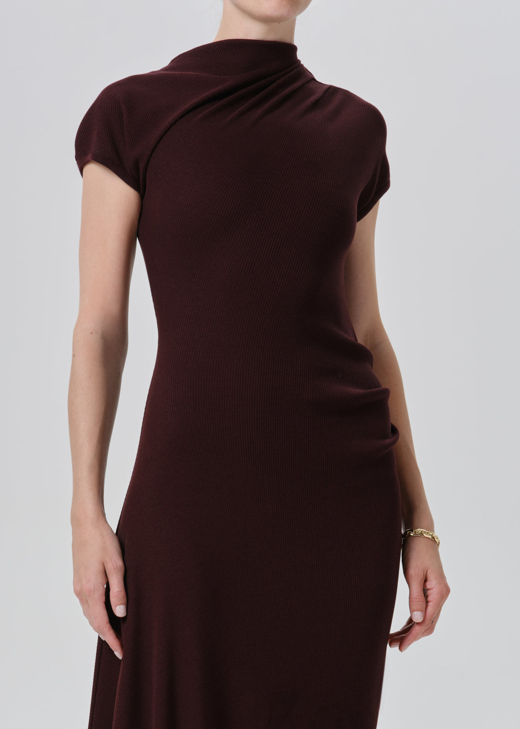 A woman wearing a dark burgundy knit dress with a draped mock neck and short cap sleeves, shown from the back against a grey background.