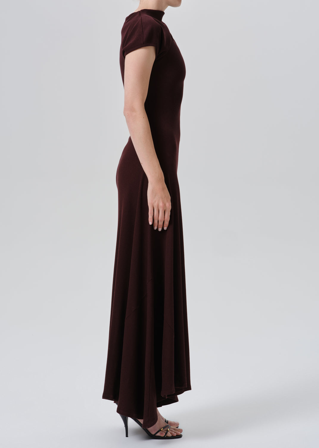 A woman modeling a dark burgundy maxi dress with short sleeves, a mock neck, and a fitted silhouette, shown in profile against a grey background.