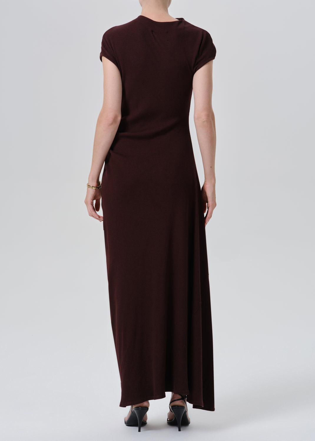 A woman viewed from the back wearing a dark brown short-sleeve maxi dress and black heels, standing against a grey background.