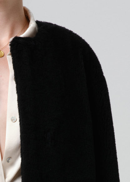 Close-up of a person wearing a black fuzzy cropped jacket or cardigan over a cream satin button-front shirt, against a grey background.