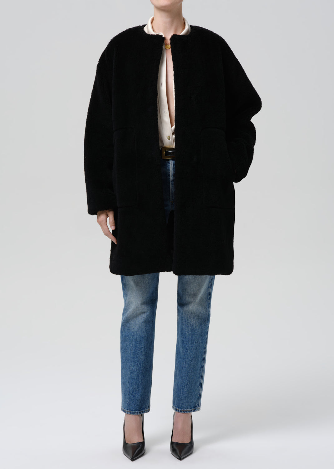 A model wearing a black oversized teddy coat over a cream blouse, blue jeans, and black pointed heels, photographed against a grey background.